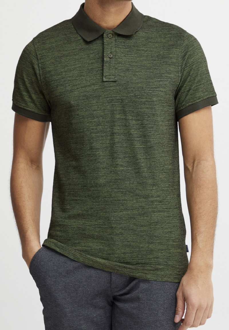 Homme portant un polo vert texturé à manches courtes avec un col vert foncé et un pantalon gris, debout avec une main dans la poche.