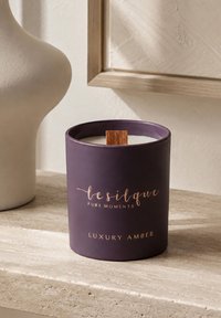 Bougie parfumée ambrée violette de luxe avec une mèche en bois sur une surface en pierre beige à côté d’un vase blanc et d’une œuvre murale encadrée.