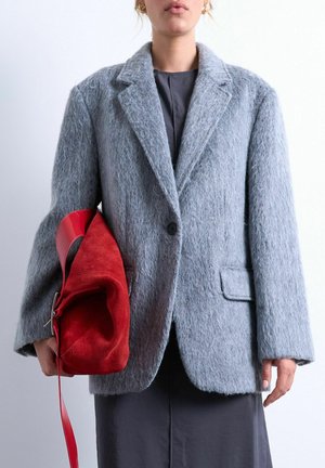 Femme portant un blazer gris texturé sur une robe noire, tenant un grand sac à main en daim rouge avec une large bandoulière en cuir dans sa main gauche.