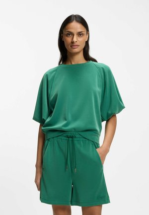 Kobieta ubrana w luźną zieloną bluzkę z krótkim rękawem i pasujące szorty ze ściągaczem, stojąca z jedną ręką w kieszeni na białym tle.