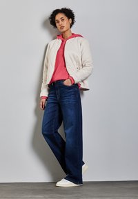 Chaqueta de crema acolchada, superpuesta sobre una sudadera rosa, combinada con unos vaqueros de pierna ancha azul oscuro y zapatillas blancas sobre un fondo gris.