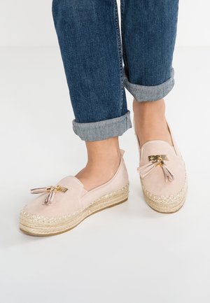 Personne portant un jean bleu retroussé et des mocassins espadrilles rose clair avec des glands et des semelles compensées tissées sur un sol blanc.
