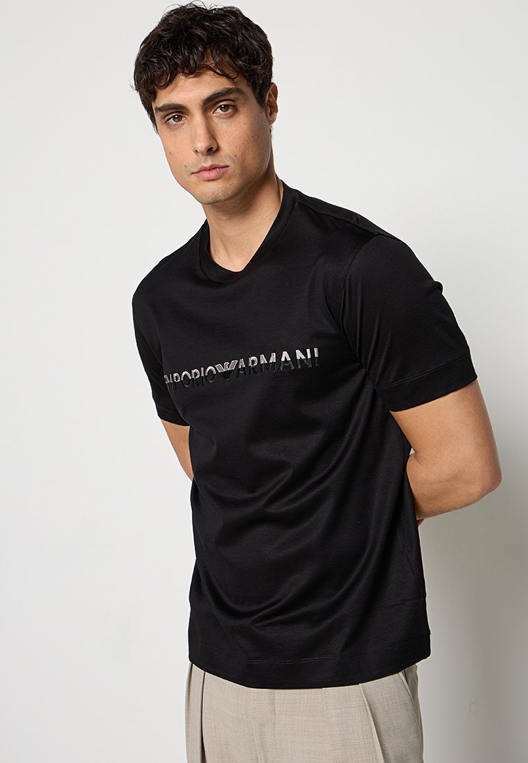 Joven con cabello rizado oscuro que lleva una camiseta negra de Emporio Armani y pantalones beige, posando frente a un fondo liso de color gris claro.