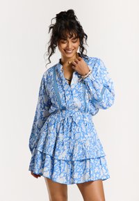 Robe bleu à motifs avec des manches longues, décolleté en v, jupe à volants en étages et taille à cordon. Design floral blanc sur un fond bleu vif.
