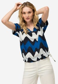 Maglietta a maniche corte e vestibilità ampia con scollatura a V. Il tessuto presenta un motivo a zigzag blu, nero e bianco. L'orlo con cordoncino aggiunge un tocco di dettaglio.