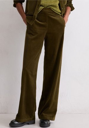 Persona vistiendo pantalones de pana de pierna ancha en color verde oliva, con las manos en los bolsillos y zapatos plataforma negros, frente a una pared lisa de color claro.