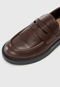 Mocassins en cuir marron au design élégant, avec une sangle sur le dessus et une finition texturée. Semelle en caoutchouc noire pour une meilleure adhérence.