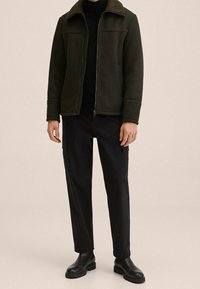 Giacca verde scuro con colletto in shearling marrone, chiusura con zip e due tasche frontali, abbinata a pantaloni cargo neri e stivali neri.