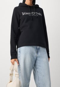 Sudadera negra de algodón con cordones, con texto de logo en blanco. Combinada con vaqueros desgastados en azul claro y un bolso crema.