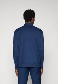 Námořnická modrá polokošile s dlouhým rukávem, plochým límcem, strukturovanou tkaninou, volným střihem a žebrovanými manžetami. Viditelné švy a minimalistický design.