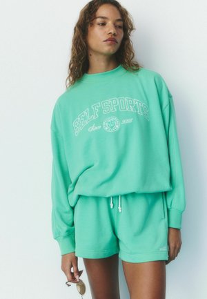 Frau trägt mintgrünes Sweatshirt mit Aufschrift "SELF SPORTS" und passende Shorts, hält eine runde Sonnenbrille in einer Hand vor weißem Hintergrund.