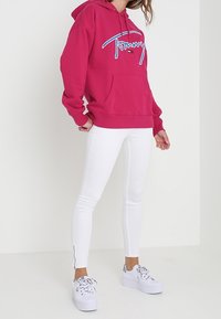 Felpa fucsia con tasca frontale e logo, abbinata a jeans skinny bianchi con cerniere laterali e sneakers bianche con dettagli neri.