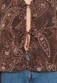 Rjav cvetlični vrh z globokim V-izrezom, zavezovanjem in zapletenimi paisley vzorci. Narejen iz lahkega blaga z teksturiranimi detajli.