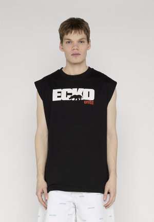 Mladić u crnoj majici bez rukava marke ECKO unltd. i bijelim kratkim hlačama s plavim natpisom ECKO unltd., stoji ispred jednobojne svijetle pozadine.