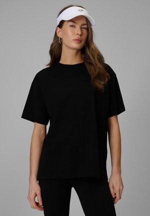 Kobieta stoi na jednolitym tle, ubrana w czarny T-shirt, czarne spodnie i biały daszek, patrzy przed siebie z neutralnym wyrazem twarzy.