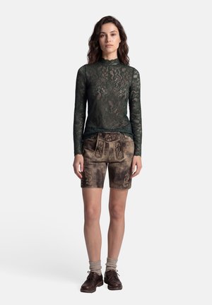 Frau steht auf weißem Hintergrund und trägt ein dunkelgrünes Spitzenoberteil mit langen Ärmeln, bestickte braune Lederhosen, beige Socken und dunkelbraune Schnürschuhe.