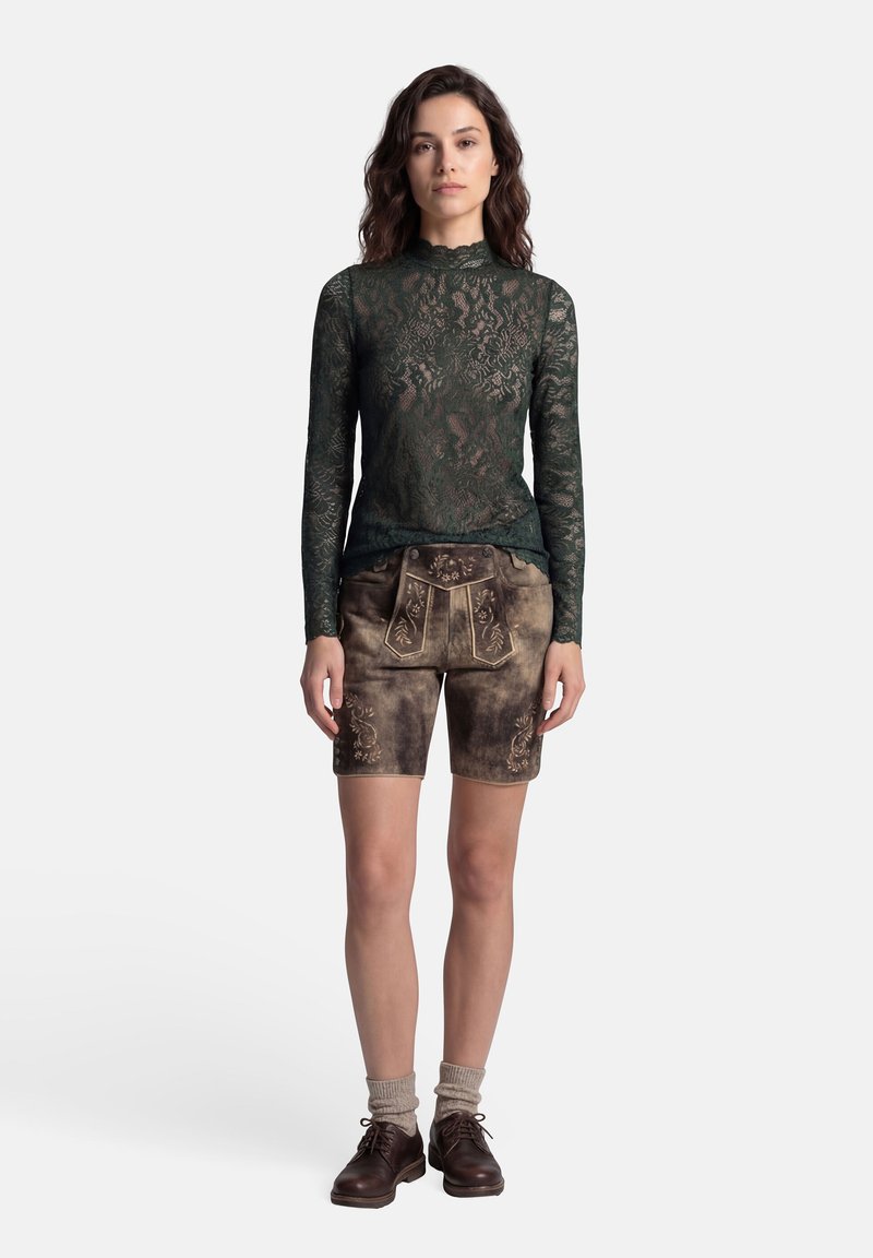 Frau steht auf weißem Hintergrund und trägt ein dunkelgrünes Spitzenoberteil mit langen Ärmeln, bestickte braune Lederhosen, beige Socken und dunkelbraune Schnürschuhe.