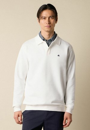 Mężczyzna ubrany w biały polo z długim rękawem na niebieską koszulę zapinaną na guziki, w granatowych spodniach, stojący na jednolitym beżowym tle.