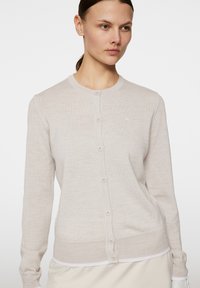 Ljusbeige cardigan gjord av ett mjukt stickat material, med rund hals, långa ärmar och knappar framtill. Subtil varumärkesdetalj på bröstet.