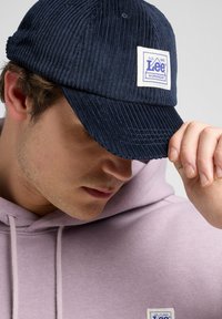 Navy corduroy cap with a curved brim and white Lee Workwear patch. Textured fabric and stitched details add to the design. Worn with a pink hoodie.