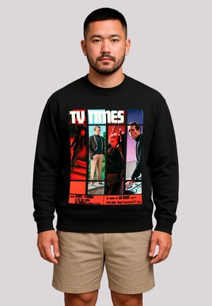 Mann trägt einen schwarzen Pullover mit einem bunten TV Times-Graphic, das vier Personen in verschiedenen Szenen zeigt, kombiniert mit beigefarbenen Shorts.