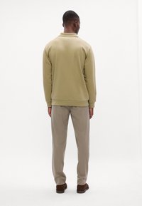 Olíva zöld pulóver gallérral, bordás mandzsettákkal és alsó résszel. Bézs nadrággal és barna cipővel párosítva, hátulról nézve.