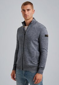 Gray zip-up sweater in a textured knit. Features a high collar and side pockets. Black zipper and logo patch on the sleeve.