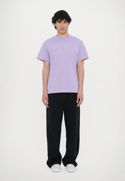 Helles lila Baumwoll-T-Shirt mit kurzen Ärmeln, Rundhalsausschnitt und minimaler Textdetail. Kombiniert mit einer lockeren schwarzen Hose und weißen Sneakern.