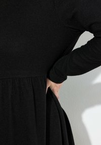 Sort, langærmet kjole lavet af tekstureret stof, snøret ind i taljen med en flydende nederdel, hånd placeret på hoften.