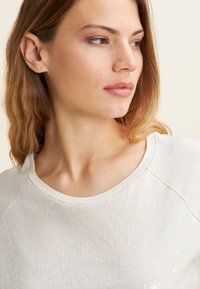 Top de color beige claro con escote redondeado, que presenta un frente texturizado y acentos bordados. Material suave con un ajuste relajado.