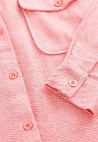 Chemise en tissu rose doux avec manches longues, poignet boutonné, poche avant et boutons roses assortis sur le devant.