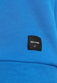 Tessuto a maglia blu con una piccola etichetta nera quadrata che recita "ONLY & SONS" e un piccolo logo a forma di cuore sotto il testo.