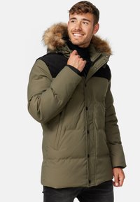Grøn polstret jakke med faux fur hætte, sorte accenter, lynlås og knaplukninger, samt et struktureret skulderområde. Inkluderer lynlåslommer.