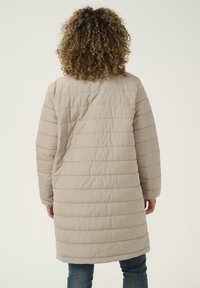Manteau matelassé beige avec des coutures horizontales, des manches longues et une coupe mi-longue. Le tissu est lisse avec une texture légèrement brillante.