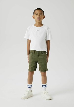 Witte t-shirt met korte mouwen en zwarte tekst, olijfgroene opgerolde shorts, witte sneakers en witte sokken met blauwe strepen.