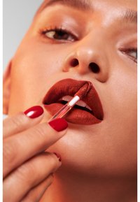 Applicazione di rossetto liquido con un applicatore trasparente su labbra di un profondo rosso marrone. Unghie dipinte di rosso brillante. Pelle liscia e sfondo neutro.