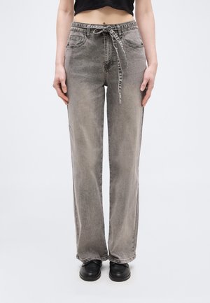 Person wearing high-waist faded gray straight-leg jeans with a tied fabric belt, black crop top, and black shoes, standing against a white background.