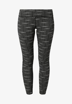 Kobieta ubrana w jasnoróżową bluzkę z długim rękawem, czarno-białe legginsy w wzory oraz czarne buty sportowe stoi na neutralnym tle.