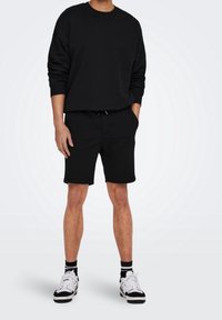 Persona che indossa una felpa nera a maniche lunghe e pantaloni corti neri abbinati, con scarpe da ginnastica bianche e nere, in piedi con le mani nelle tasche.