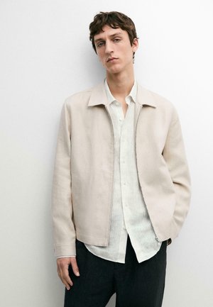 Ung mand iført en beige jakke over en lys, stribet skjorte med knapper og mørke bukser, poserer mod en ensfarvet hvid baggrund.