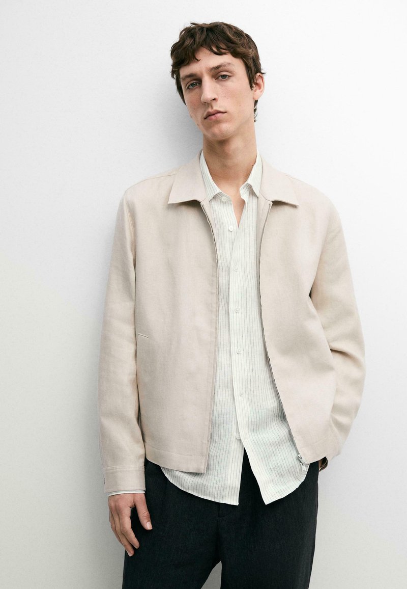 Junger Mann, der eine beige Jacke über einem hell gestreiften Hemd mit Knöpfen und dunkler Hose trägt, vor einem schlichten weißen Hintergrund posiert.