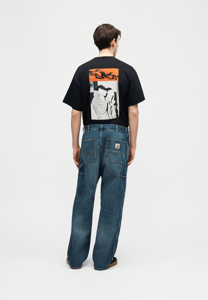 Joven de espaldas con camiseta negra con un diseño abstracto en naranja y blanco, combinada con vaqueros azules holgados y zapatos casuales.