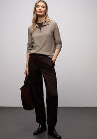 Tröja i ljusbeige ribbad tyg med dragkedja vid kragen, kombinerad med löst sittande mörkbruna byxor. Modellen håller en mörkbrun väska.