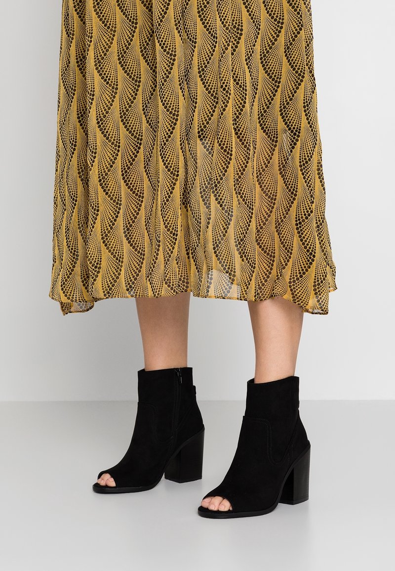 Bottines en daim noir à bout ouvert avec des talons carrés, portées avec une jupe midi à motif jaune moutarde.
