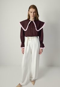 Bordeaux blouse met een grote gerimpelde kraag in roze, knoopsluiting aan de voorkant, lange mouwen met gerimpelde manchetten, gecombineerd met witte wijde broekspijpen.