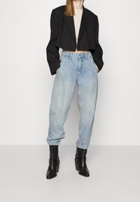 Lyhyttoppinen musta bleiseri, beige paita, vaaleansiniset korkeavyötäröiset farkut ja mustat korkokengät solkiyksityiskohdilla. Silkkinen kangas, rento istuvuus.