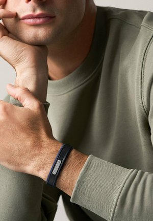 Mann ruht mit dem Kinn auf der Hand, trägt ein olivgrünes Sweatshirt und ein schwarzes Lederarmband mit einem metallischen Fossil-Logo.