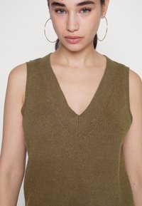 Sweater sem mangas verde azeitona com um decote em V profundo, textura canelada e detalhes subtis na gola. Tecido de malha suave.