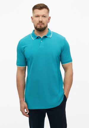 Man met baard die een groenblauw poloshirt met korte mouwen en witte bies draagt, staand met één hand in zijn zak tegen een effen witte achtergrond.