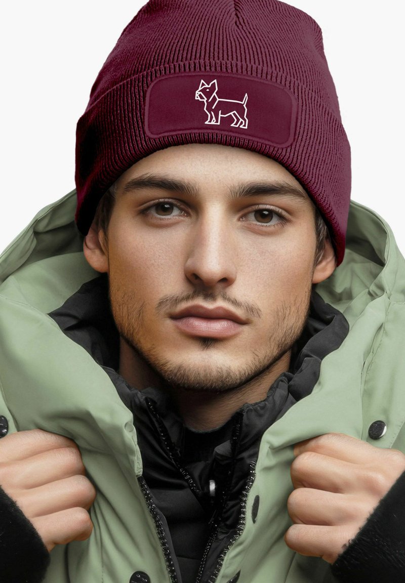 Maroon knit beanie with a ribbed texture and a stitched white dog silhouette on a contrasting patch; worn with a green puffer jacket.